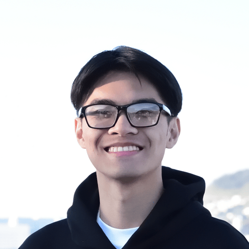 A young man with short dark hair and glasses smiles at the camera, wearing a black hoodie outdoors with a bright sky in the background.