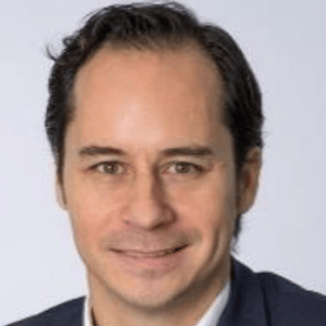 A man with short dark hair and a slight smile is wearing a dark jacket and light shirt against a plain light background.