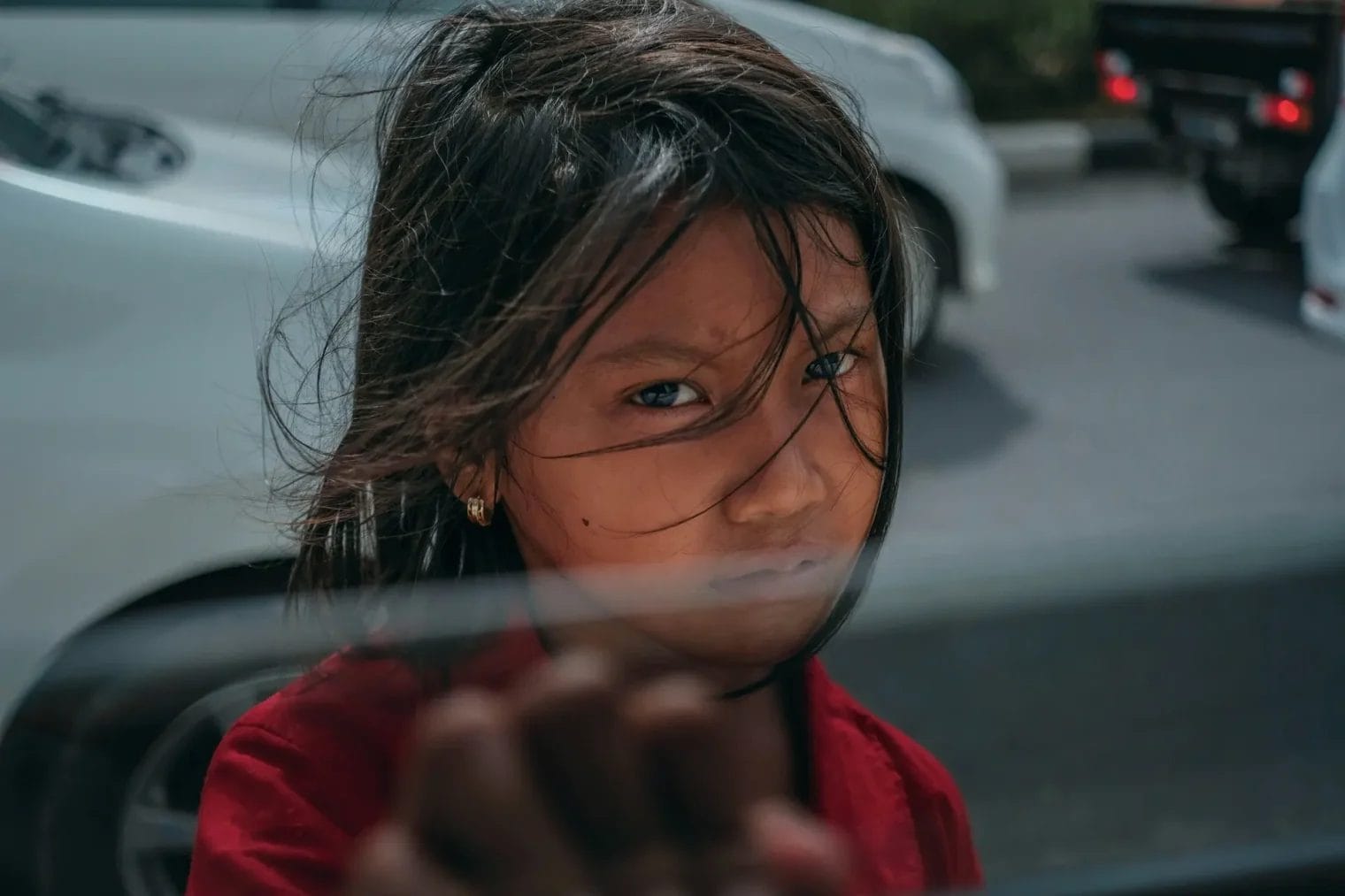 A young girl with long hair stands outside by a road, looking through a glass window with her hand raised, with cars visible in the background.