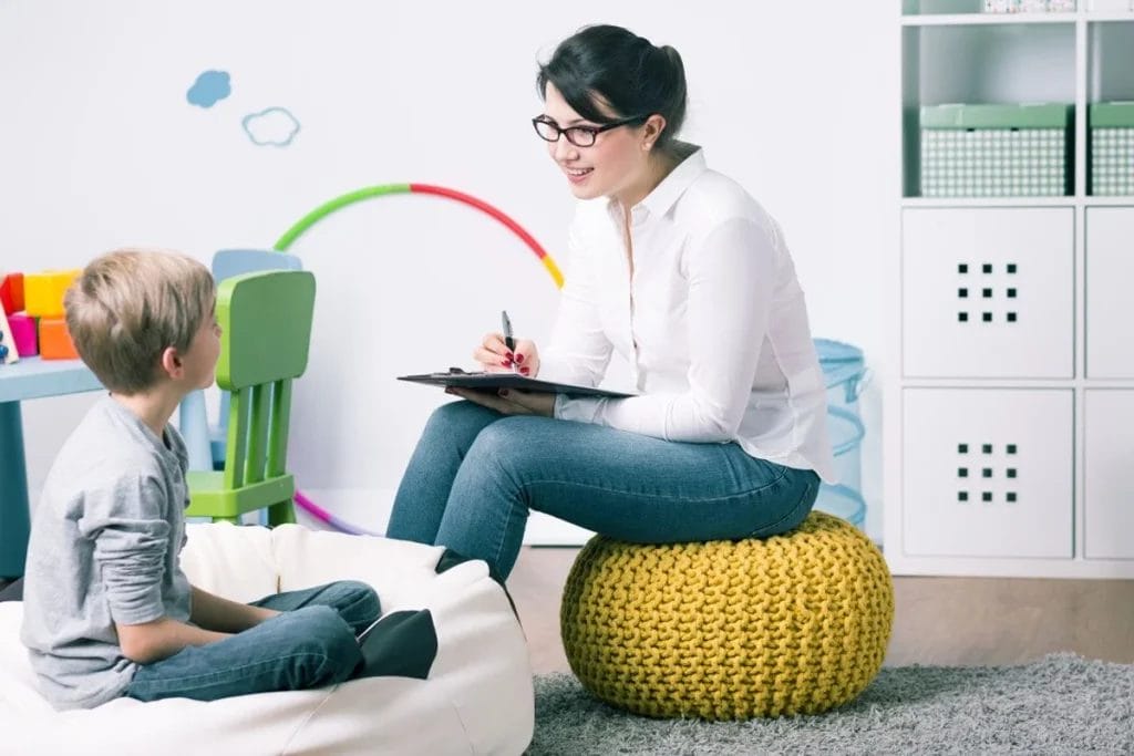 A woman with a clipboard speaks to a seated boy in a brightly lit, child-friendly room with toys and colorful decor.
