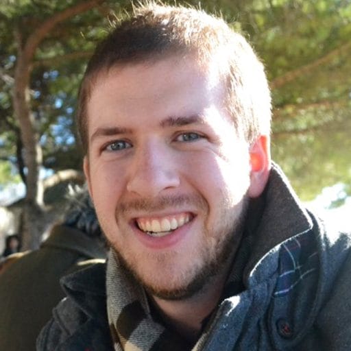 A man with short hair and a beard is smiling outdoors, wearing a scarf and a jacket. Trees and sunlight are visible in the background.