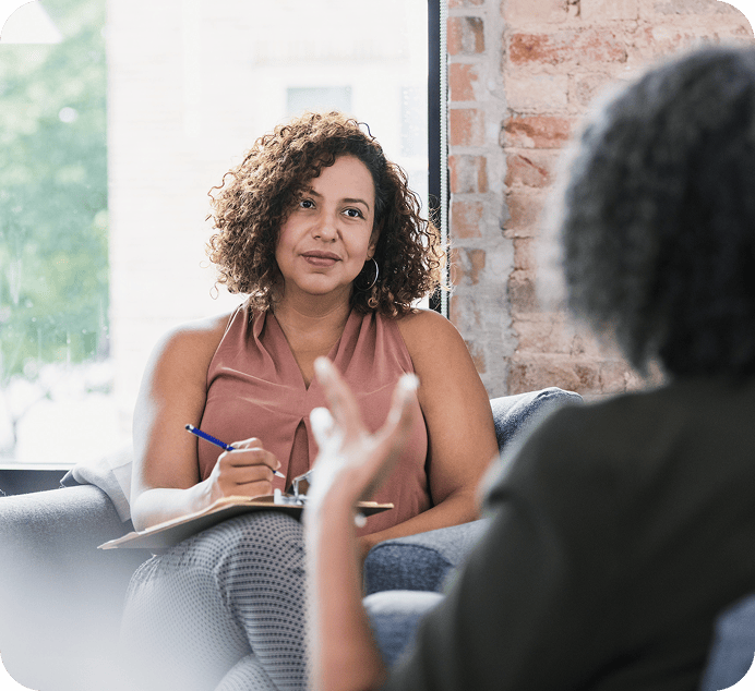 A woman holding a clipboard and pen listens attentively to another person speaking, eager to explore new solutions during their conversation in a bright, modern room.