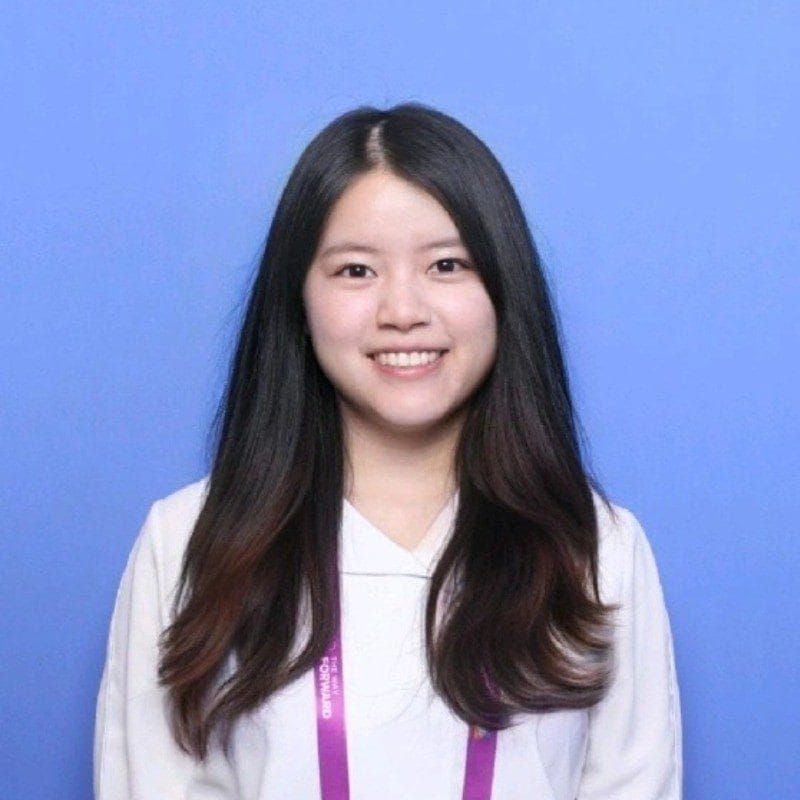 A woman with long dark hair, wearing a white top and a purple lanyard, smiles at the camera against a plain blue background.