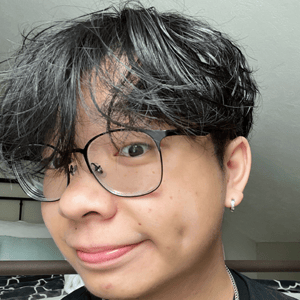 A person with black hair and glasses smiles slightly, showing dimples. They are indoors with a bed and a white wall visible in the background.
