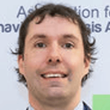 A person with wavy brown hair and a light complexion is smiling at the camera in front of a blurred sign with partially visible text.