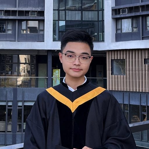 A person wearing glasses and a black and yellow graduation gown stands outside a building with large windows.