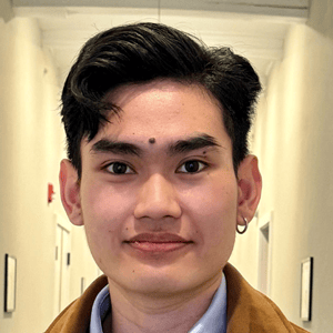 A young person with short black hair, wearing a brown jacket, blue shirt, and a small hoop earring, stands in a hallway with white walls and doorways visible in the background.