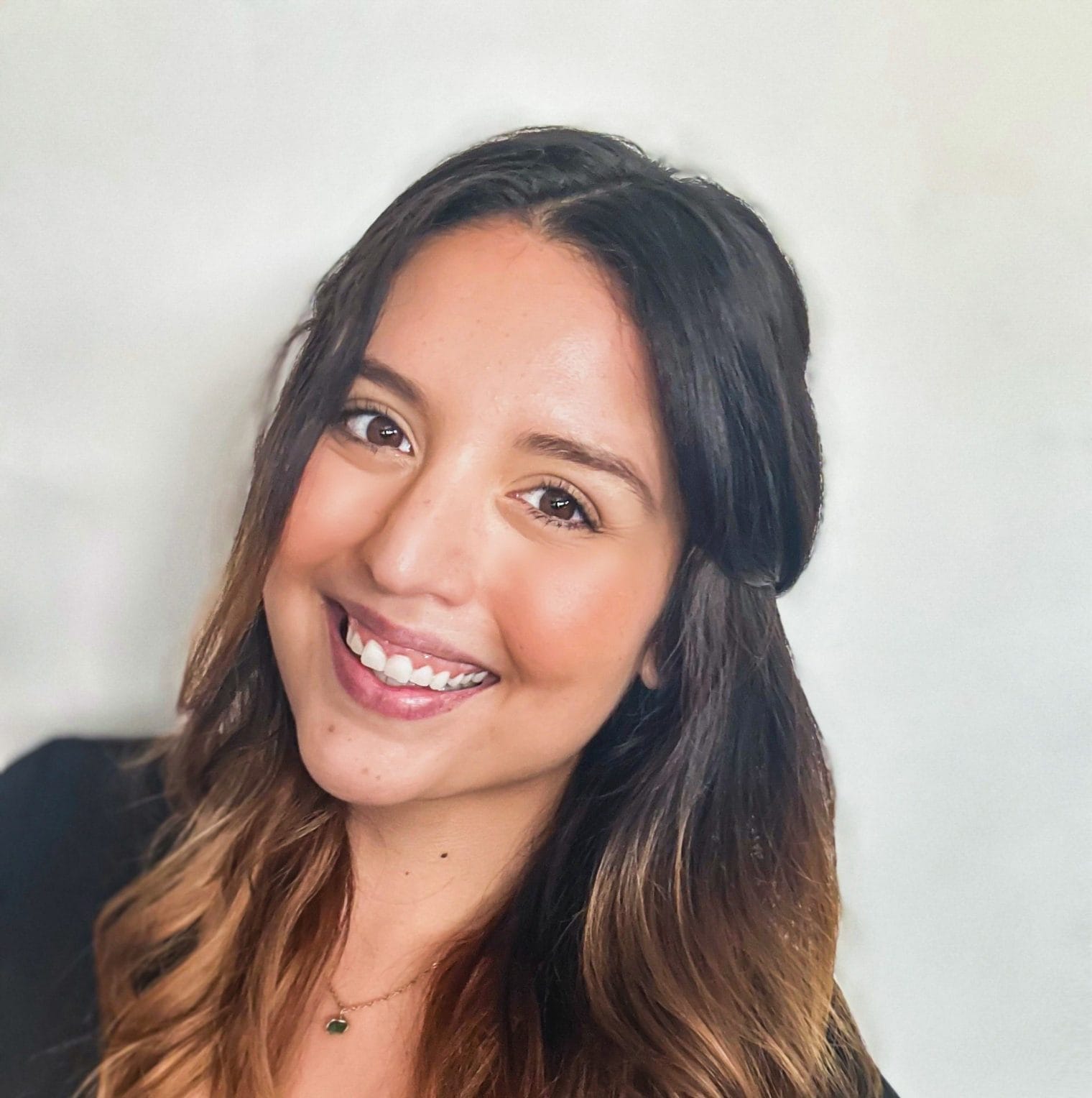 A woman with long brown hair and a black top smiles at the camera against a plain light background.