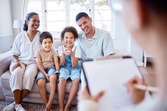 A family of four sits on a couch, smiling, while a person in the foreground takes notes on a clipboard.