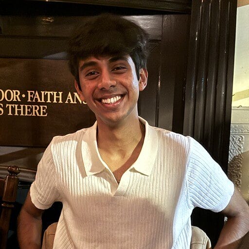 A young man with dark hair smiles at the camera, wearing a white short-sleeved shirt and sitting indoors in front of a wall with partially visible text.