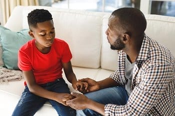 A man sits on a couch holding hands with a boy in a red shirt, appearing to have a serious conversation about effective interventions for cyberbullying.