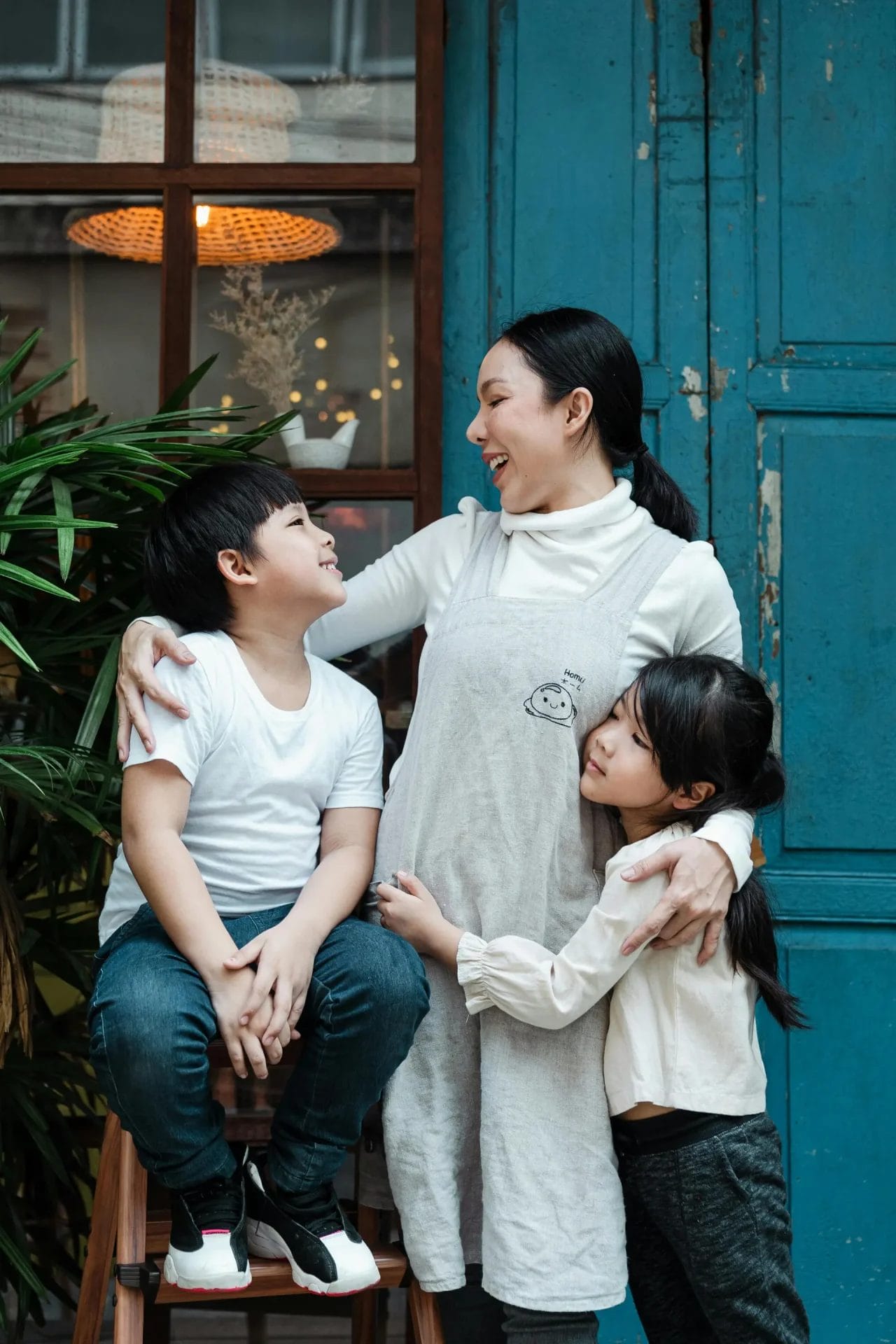 A woman stands smiling with her arms around a young boy and girl, all looking at each other, in front of a blue door and plants.