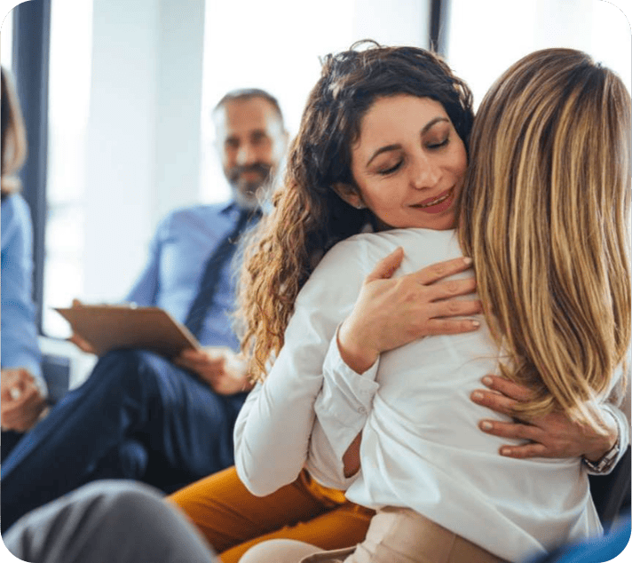 Two women are hugging each other in an indoor setting, while a man with a clipboard sits in the background, suggesting a support or therapy group environment.