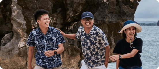 Three people wearing patterned shirts and hats smile and laugh together while standing by rocky cliffs near the ocean.