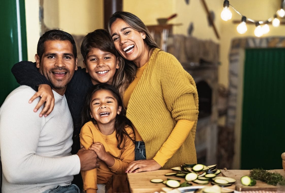A family of four smiles and hugs together in a kitchen, with sliced zucchini on the counter in front of them.