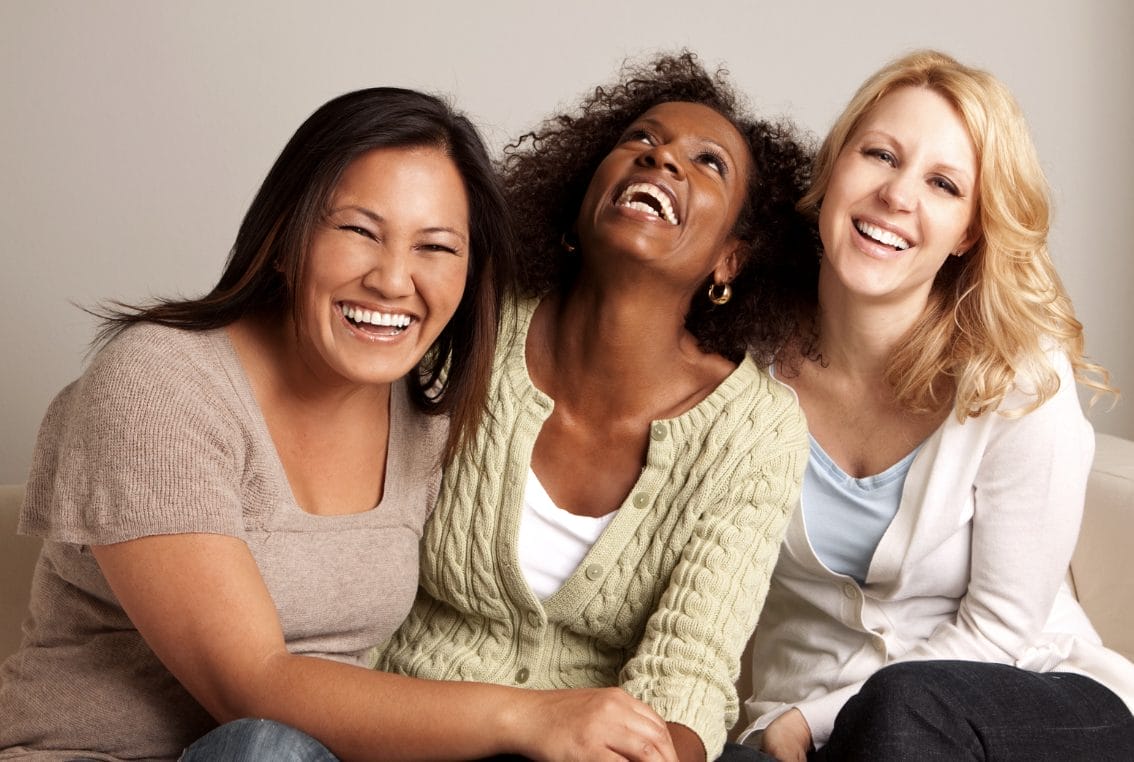 Three women sit closely together on a couch, smiling and laughing, in a well-lit indoor setting.