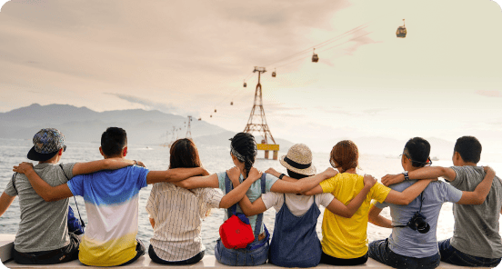 Seven people sit side by side on a ledge by the water, arms around each other, facing away, with cable cars visible in the background over the sea.