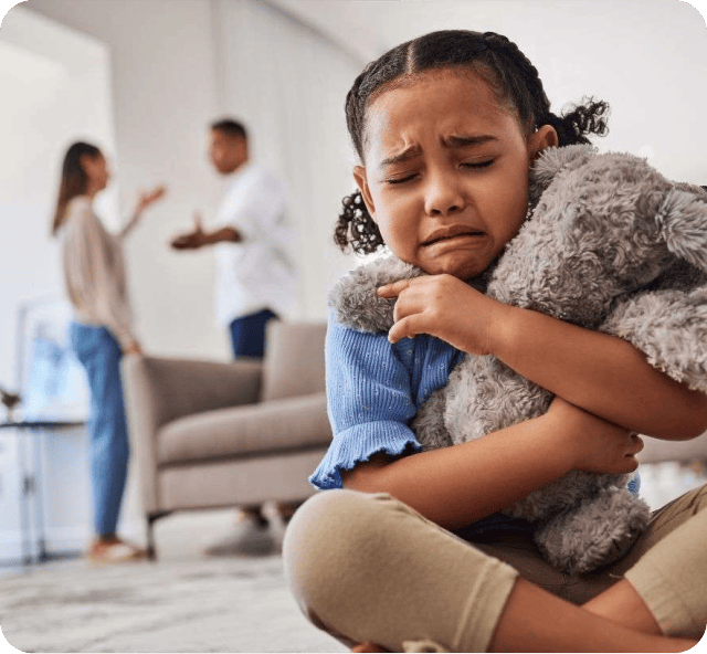 A young girl hugs a stuffed animal and cries while two adults argue in the background of a living room.