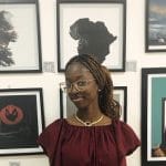 A woman wearing glasses and a maroon top stands in front of framed artwork on a white wall, reflecting on the effects of cyberbullying depicted in the display.