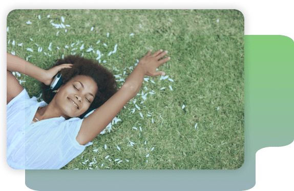 A person lies on grass with petals around them, eyes closed and smiling, wearing headphones and a white shirt.